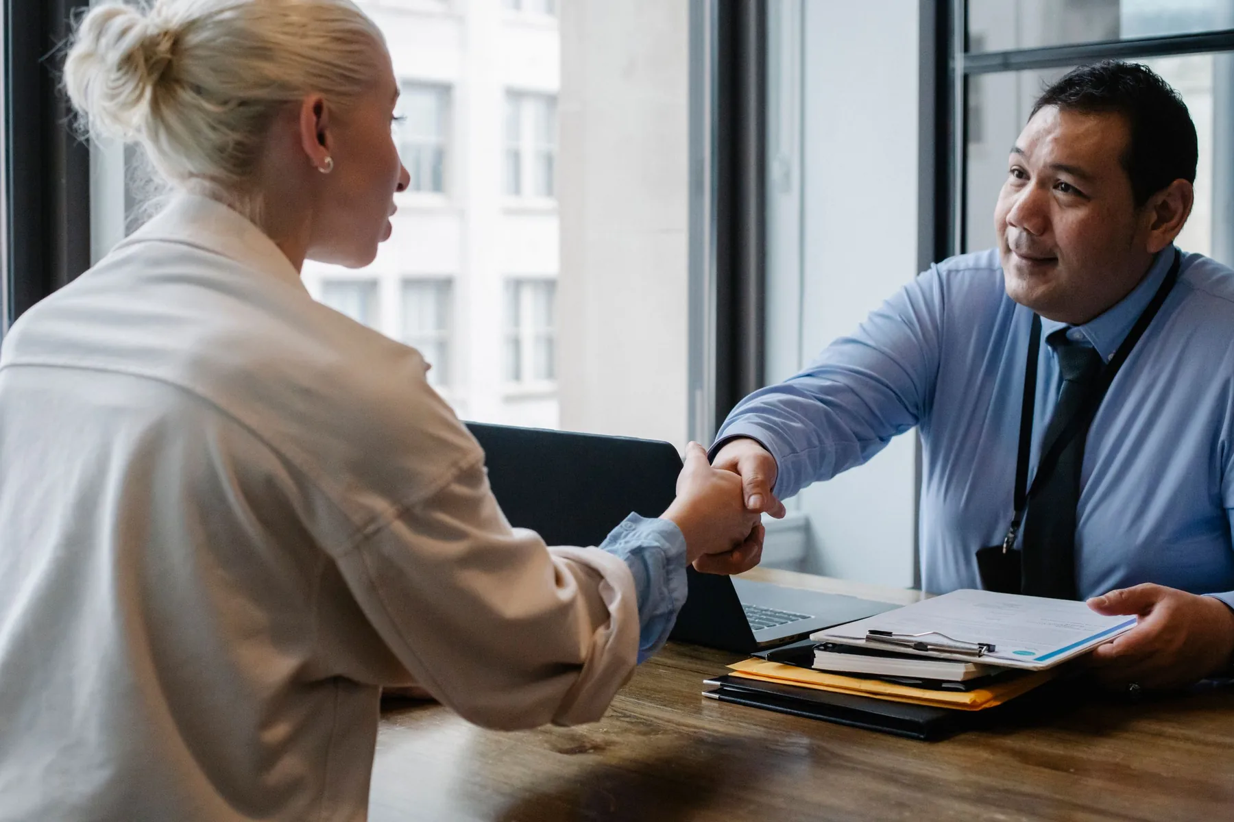 Two people shaking hands across a desk beside large windows